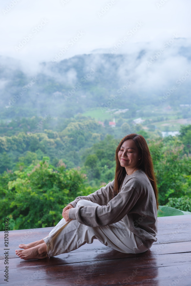 Fototapeta premium Vertical image of a woman sitting on wooden balcony with a beautiful mountain views on cloudy morning