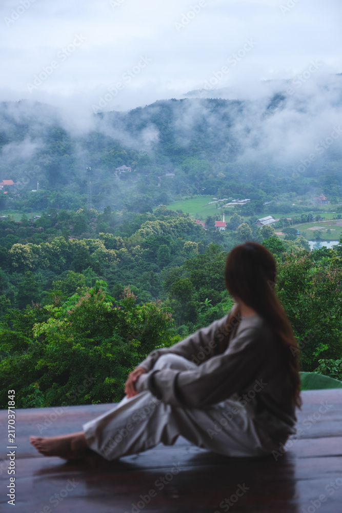 Fototapeta premium Vertical rear view image of a woman sitting on wooden balcony with a beautiful mountain views on cloudy morning