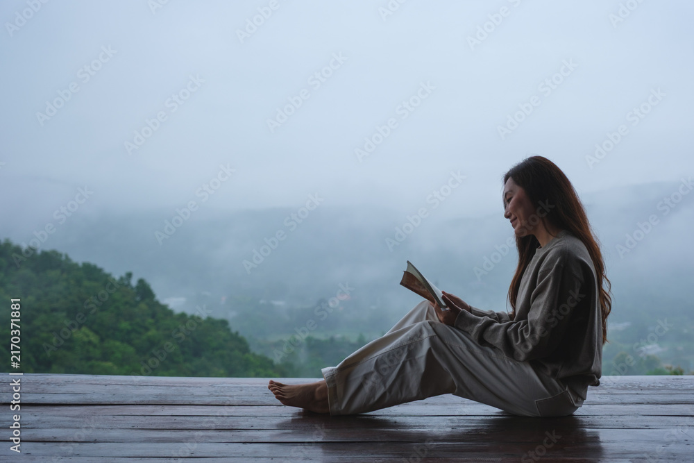Fototapeta premium A woman reading a book while sitting on wooden balcony with a beautiful mountain views on cloudy morning