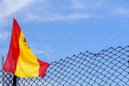 Spanish flag hanging on a chain link fence against a bright blue sky, symbolizing borders, restrictions, and national identity