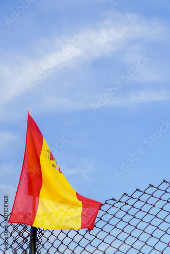 Spanish flag with national coat of arms waving on a metal fence. Clear blue sky with light clouds in background