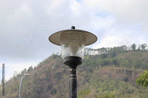 A close-up of a modern outdoor street lamp post set against a backdrop of cloudy sky and misty, forested green mountains.