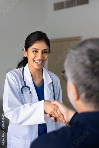 Smiling South Asian female doctor in white coat shaking hand, greeting male patient in clinic office. Trust in healthcare services, professional consultation, positive outcome of medical visit concept