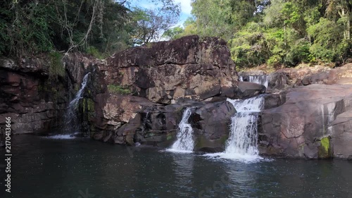 Several small waterfalls cascade over a rugged rock formation into a dark natural pool below.