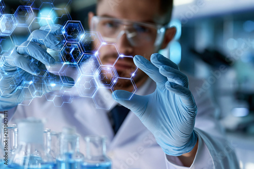Scientist holds glowing molecular structure in lab. Hands gesture toward intricate hexagon patterns. Blue gloves and safety glasses complete lab attire. Background shows glassware  blurred equipment