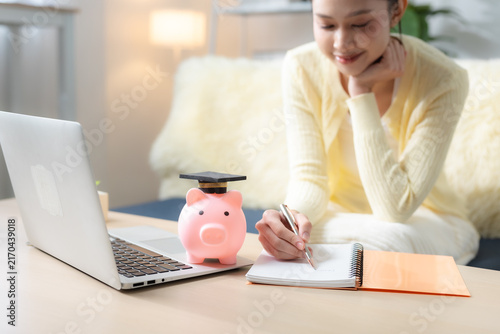 Фотография Woman planning education savings with piggy bank in graduation cap, laptop and notebook at home, representing college fund, student finance, budgeting and future education goals