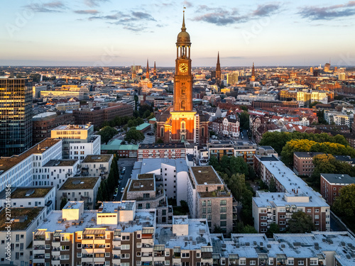 Aerial view of St. Michaeliskirche illuminated by golden hour light, surrounded by historic rooftops and the urban skyline of Hamburg, Germany.
