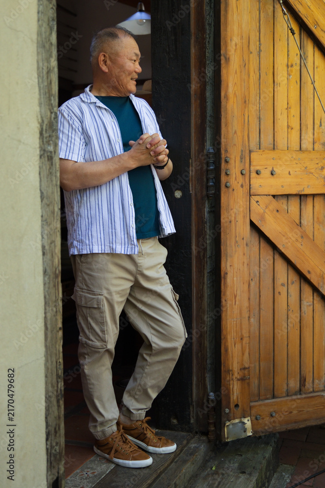 Fototapeta premium Senior Asian man standing on threshold in striped tee, leaning against planked door, clasping hands