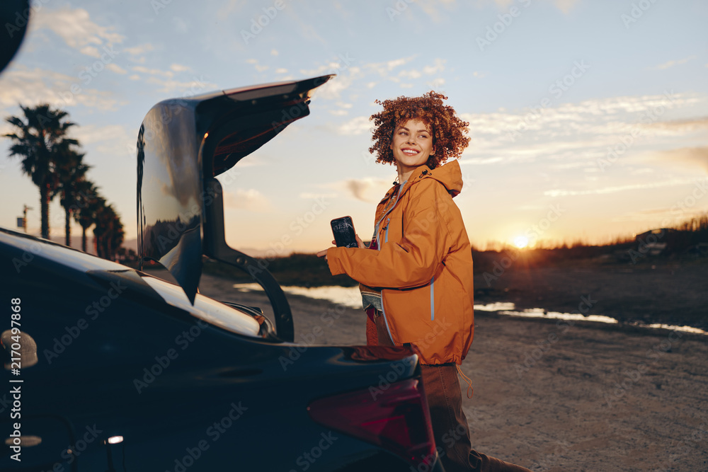 Fototapeta premium Woman lifestyle near open car trunk at sunset, curly hair portrait, holding smartphone, relaxed smile, warm golden light, casual jacket, outdoors travel mood, soft breeze, natural colors
