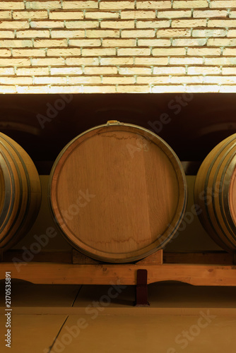 Obraz na płótnie Wooden oak wine barrel stored in a winery cellar, captured under warm light with a brick wall background, representing traditional winemaking and the wine aging process