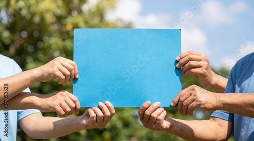 Photorealistic close-up of diverse Asian hands holding a blank blue environmental sign outdoors, with blue sky and green trees softly blurred behind, natural sunlight and Clean Air Day symbolism.