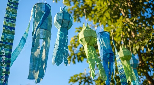 Photorealistic close-up of blue and green eco-friendly decorations moving in the breeze, framed by a clear blue sky and green leaves, with soft sunlight and elegant minimal Clean Air Day styling
