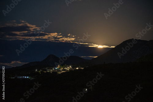 Photography The moon begins its ascent over a mountain, its brilliant light hidden behind a cloud, illuminating a tranquil Greek village nestled in the dark valley below