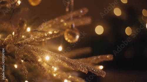 A close-up of a Christmas tree branch coated in shimmering glitter, tiny reflective particles catching and scattering warm golden light as the camera slowly moves past, delicate ornaments
