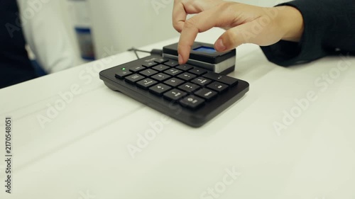 Close-up of person's hand typing PIN code on black numeric keypad payment terminal on white table. Business finance and payment concept for banking, retail, technology and commercial advertising