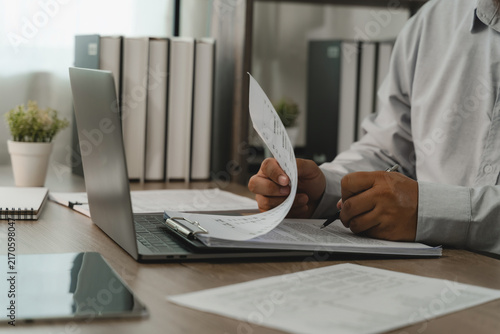 Businessman checking payroll documents on clipboard beside laptop for payroll processing, salary review, employee records, HR administration, accounting workflow.