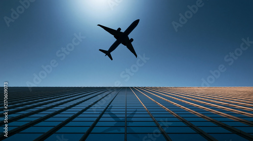 Silhouette of an airplane flying low over a modern skyscraper with reflective glass windows under a bright sun. Perfect for travel and business aviation concepts.