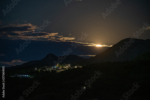 Принти на полотні The bright full moon begins to crest a mountain peak, its light spilling over the silhouetted landscape and a quiet, illuminated Greek village at night
