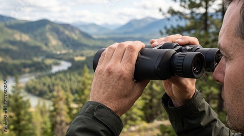 Middle-aged man using binoculars to look at a scenic mountain landscape and winding river in the wilderness during daytime for outdoor exploration and wildlife viewing
