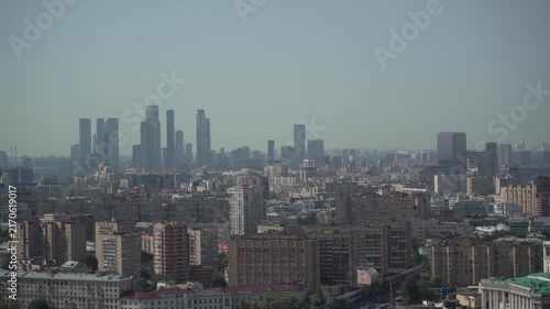 Panoramic elevated cityscape showing dense lowrise foreground and distant modern highrise skyline under hazy daylight, with cityscape, skyline, skyscraper highrise detail
