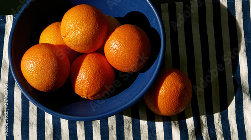 Fresh oranges in blue ceramic bowl on striped fabric, ripe citrus fruit in natural sunlight top view