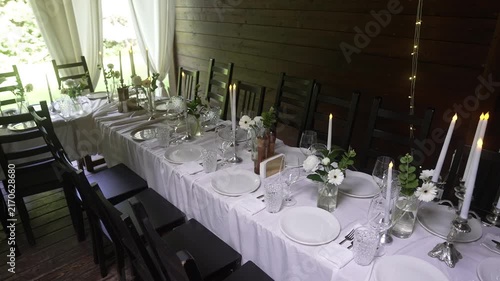 Long rustic banquet table set with white linens, plates, crystal glassware, silver candlesticks, and fresh white floral centerpieces on covered wooden porch leading toward