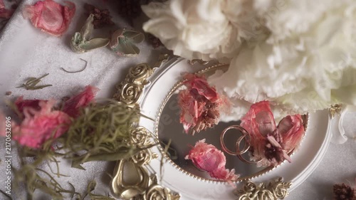 Top-down close-up of tabletop still life with white ruffled bloom, scattered pink petals, ornate gilded mirror, and gold rings in elegant vintage styling, with mirror,ring, petal, flower detail