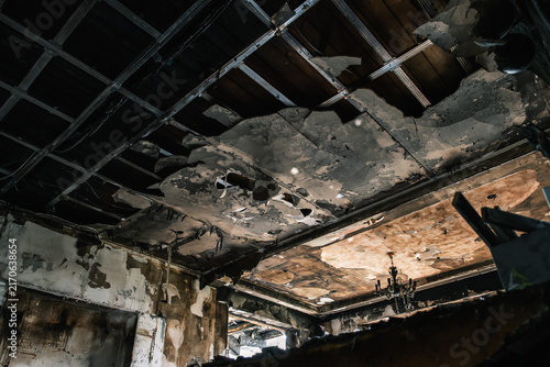 A room interior shows a heavily fire damaged ceiling with an exposed metal grid, missing panels, and visible charring and smoke residue, destruction, disaster, damage. Vandalism debris.