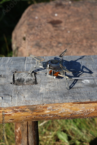 Close-up photo of a compact, portable camping stove resting on a weathered wooden beam outdoors.