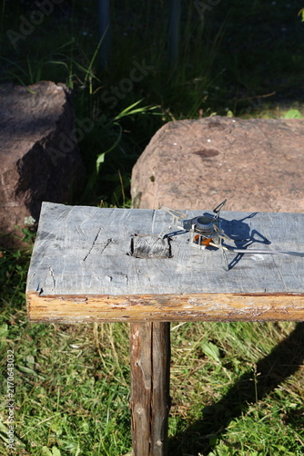 Close-up photo of a compact, portable camping stove resting on a weathered wooden beam outdoors.