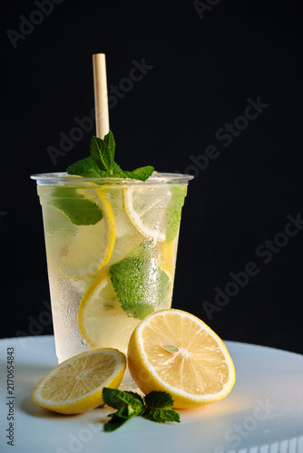 A vertical shot of a clear takeaway cup with cold lemonade, ice, and mint next to fresh lemon halves on a dark background for beverage advertising and menu campaigns.