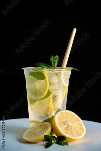 A vertical shot of a clear takeaway cup with cold lemonade, ice, and mint next to fresh lemon halves on a dark background for beverage advertising and menu campaigns.