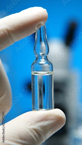 Laboratory technician in white gloves holds a clear glass ampoule filled with liquid, showcasing the ampoule against a blue background with a microscope in view