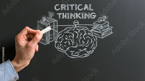 Human hand pointing to books and a brain below critical thinking text on blackboard, ideal for education, problem solving and analytical learning.