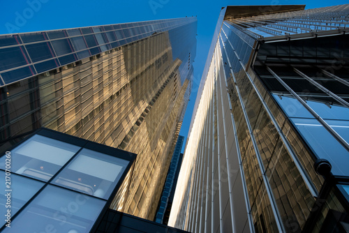 Upward view between modern towers in London within the United Kingdom with reflective glass facades and vertical lines against blue sky emphasizing urban height