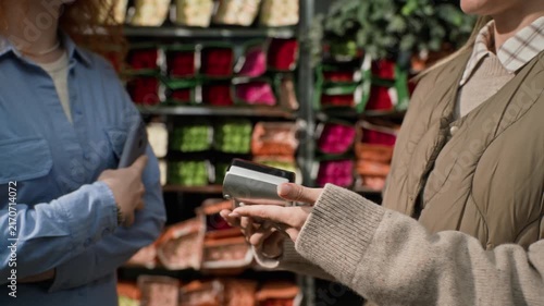 Caucasian female customer paying with smartphone via NFC on payment terminal held by female florist in flower warehouse. Perfect for retail technology and contactless payment marketing campaigns