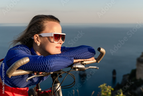 Принти на полотні Cyclist, woman, sea, resting on her bicycle handlebars at golden hour while look