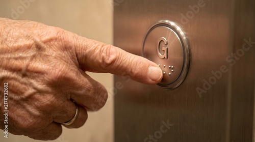 Close-up of a senior person's finger pressing the ground floor elevator button featuring braille for accessibility.
