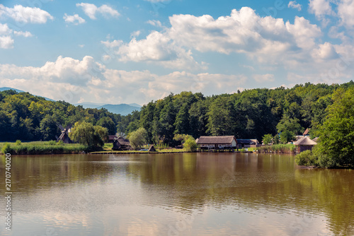 Scenic lake with traditional wooden buildings windmills forest and distant mountains under cloudy sky.
