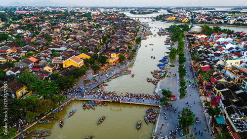 Aerial view of Hoi An Ancient Town with traditional wooden boats on the Thu Bon River, historic yellow buildings, and people walking along the waterfront in Hoi An, Da Nang, Vietnam.