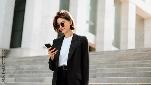A stylish woman in a black suit and sunglasses, standing outside a modern building and looking at her phone. The sun is shining brightly, highlighting her confident demeanor