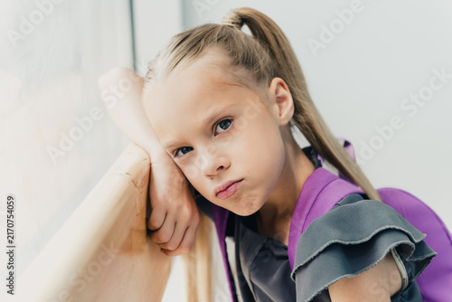 Konstfotografi Bored schoolgirl leaning on a windowsill with a purple backpack, looking frustra