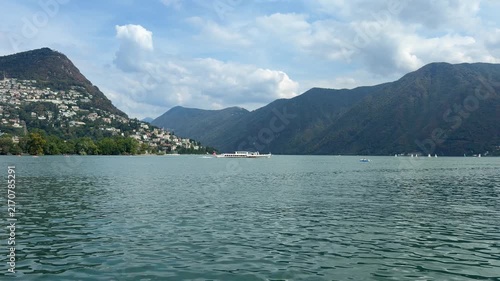 A breathtaking view of a tranquil lake nestled between majestic mountains, with a charming town visible on the hillside under a vast, partly cloudy sky. Switzerland, Lugano, Lake Lugano