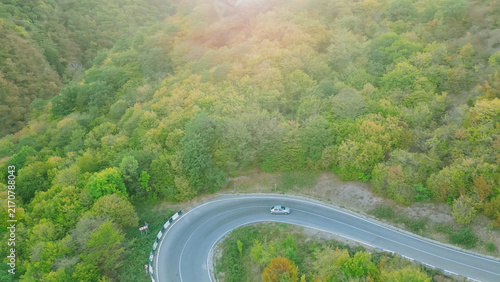 Aerial high angle view of silver car driving around hairpin curve turn on asphalt road through dense mountain forest horizontal stock photo