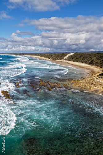 Vertical aerial view of the rugged headland and rocky coastline at Brooms Head with the long sandy shoreline of Sand Dune Beach, Pacific Ocean and surrounding natural coastal landscape 