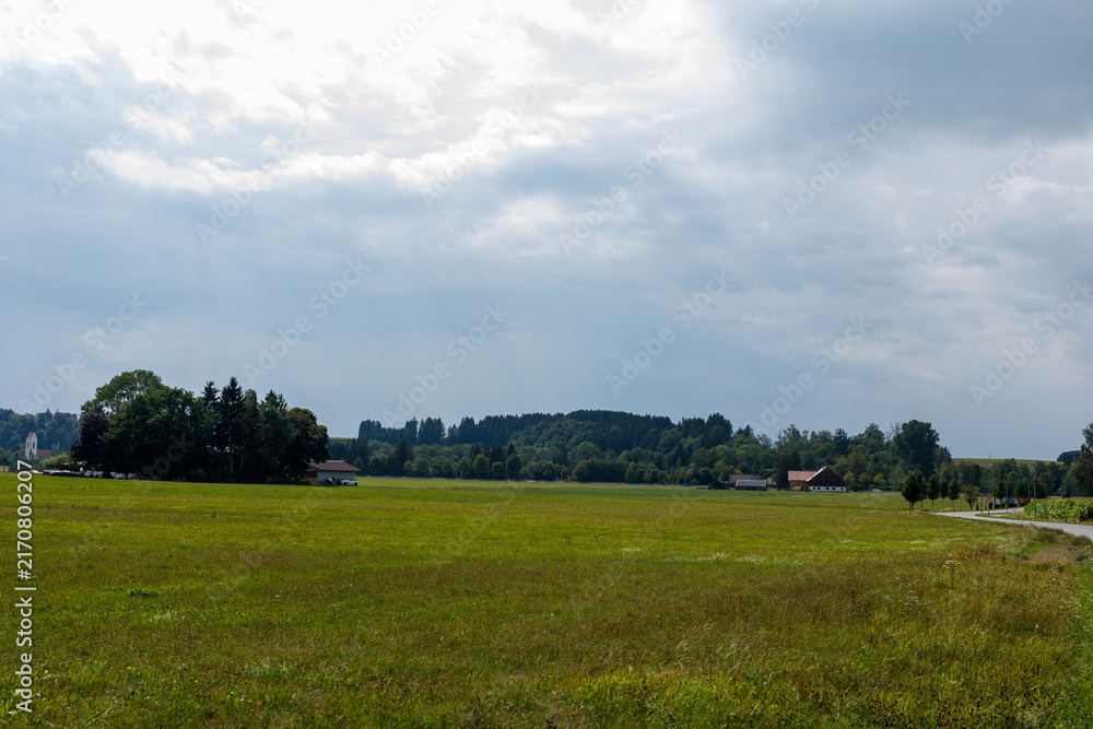 Fototapeta premium Expansive green fields under a cloudy sky with distant forests, rural buildings, and a winding road.