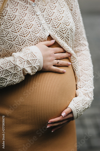 A close-up photo of a young pregnant woman in a dress, gently holding her large belly in her hands. Waiting for a miracle. Pregnancy concept.