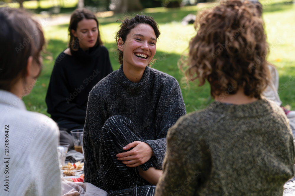 Obraz premium A cheerful woman laughing joyfully while sitting in a circle of friends on a picnic blanket, creating a warm and inviting atmosphere filled with happiness.