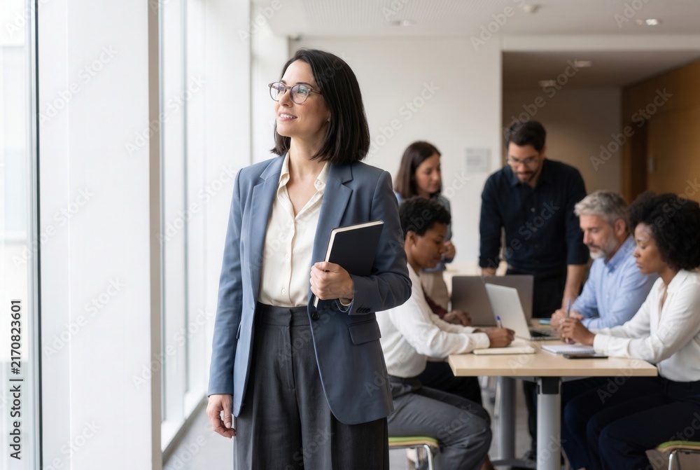 Obraz premium Confident businesswoman holding a notebook, looking at the future, with team collaborating in background