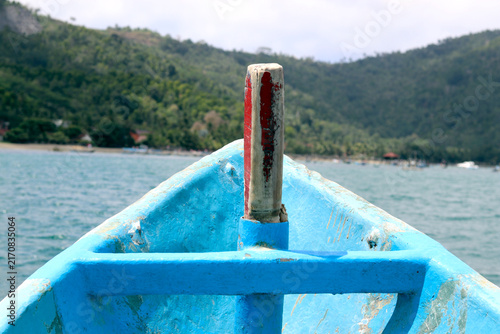 A close-up view looking over the blue wooden bow of a boat heading through calm turquoise waters toward a lush, green mountain coastline.